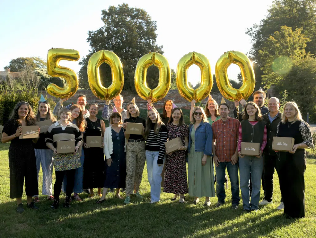 Little Lifts team celebrate delivering 50000 Little Lifts care package Boxes to breast cancer patients. They stand smiling outside, wearing party hats with gold 50000 balloons