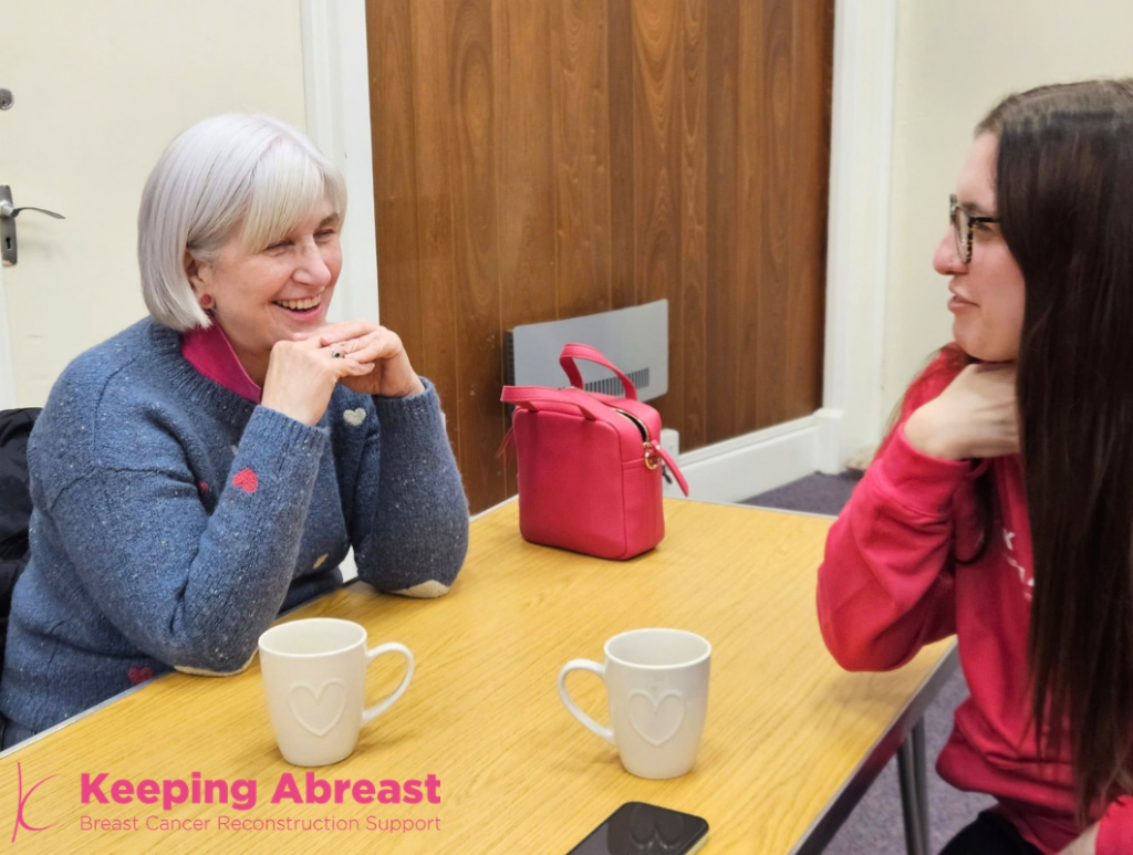 Two women sit at a table with mugs discussing breast reconstruction surgery support