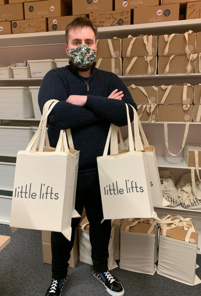 George holding several branded Little Lifts tote bags, wearing a face mask, shelves of Boxes behind