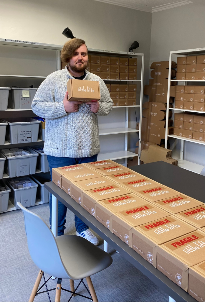 George holding a single Little Lifts Box, with rows of packed Boxes on the table in front of him