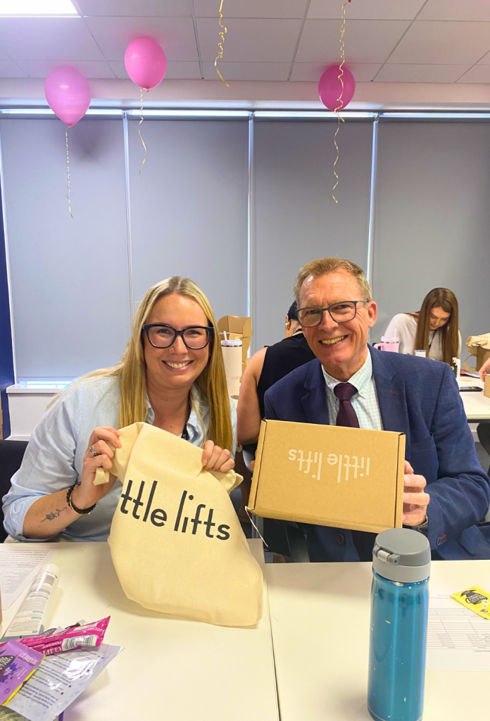 Two New Homes Group colleagues holding a Little Lifts tote bag and breast cancer care box.