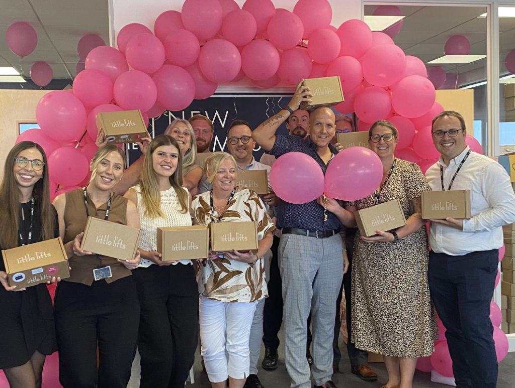 The New Homes Group team celebrating their Little Lifts Packing Party, holding Little Lifts breast cancer care boxes under a pink balloon arch.