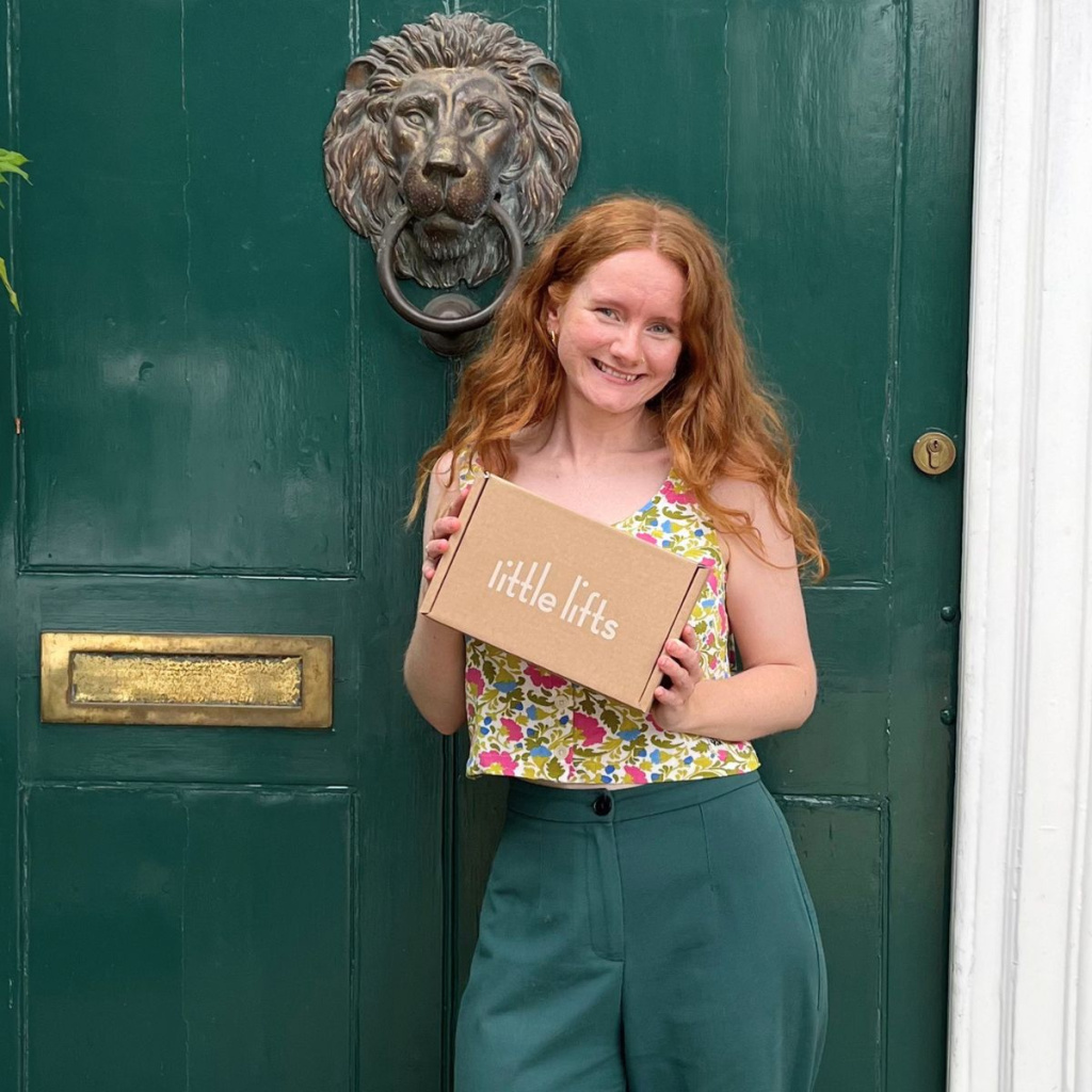 Izzy Johnson standing outside the front door to Little Lifts HQ, holding a Little Lifts Box.