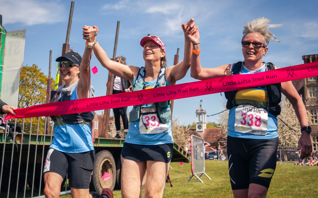Half marathon trail runners cross the Little Lifts trail run finish line holding hands and running through a pink little lifts branded ribbon